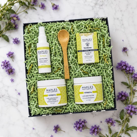 A flat-lay of the Verbena Luxe Box with green shredded paper. The set includes a body oil spray, a wooden spoon, a soap bar, a body salt, and a body butter, all with yellow and white packaging, on a white marble surface with purple flowers scattered around.